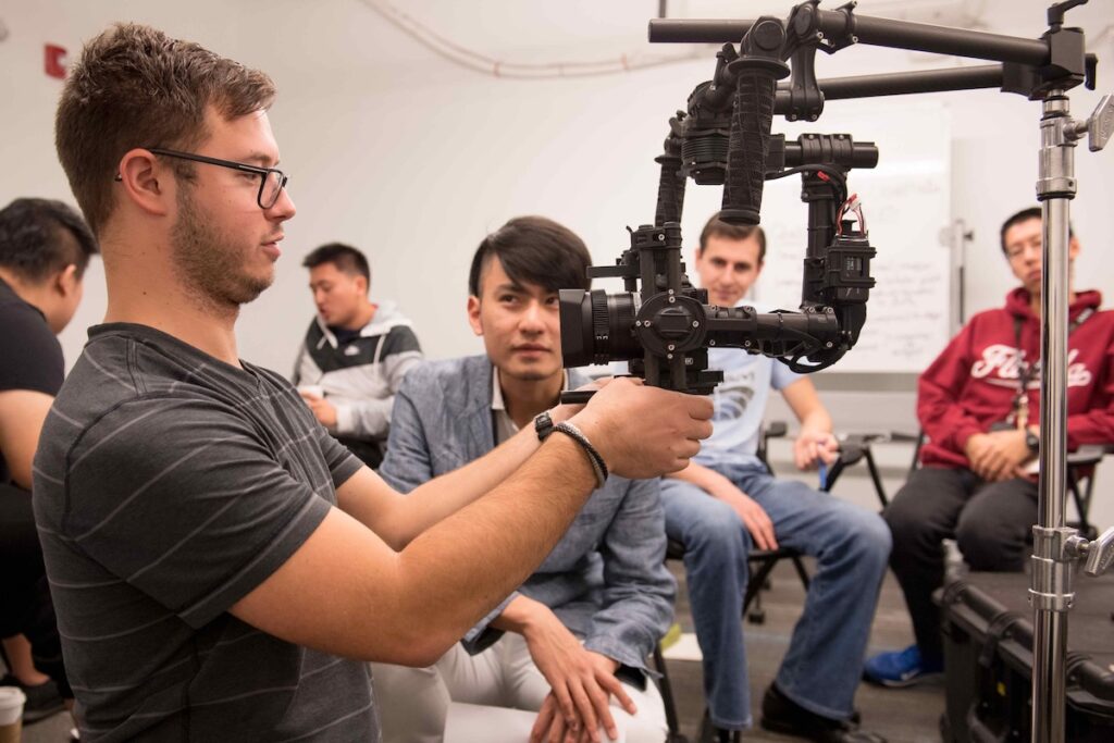 A small room of Emerson College film students being shown how to use a piece of camera equipment