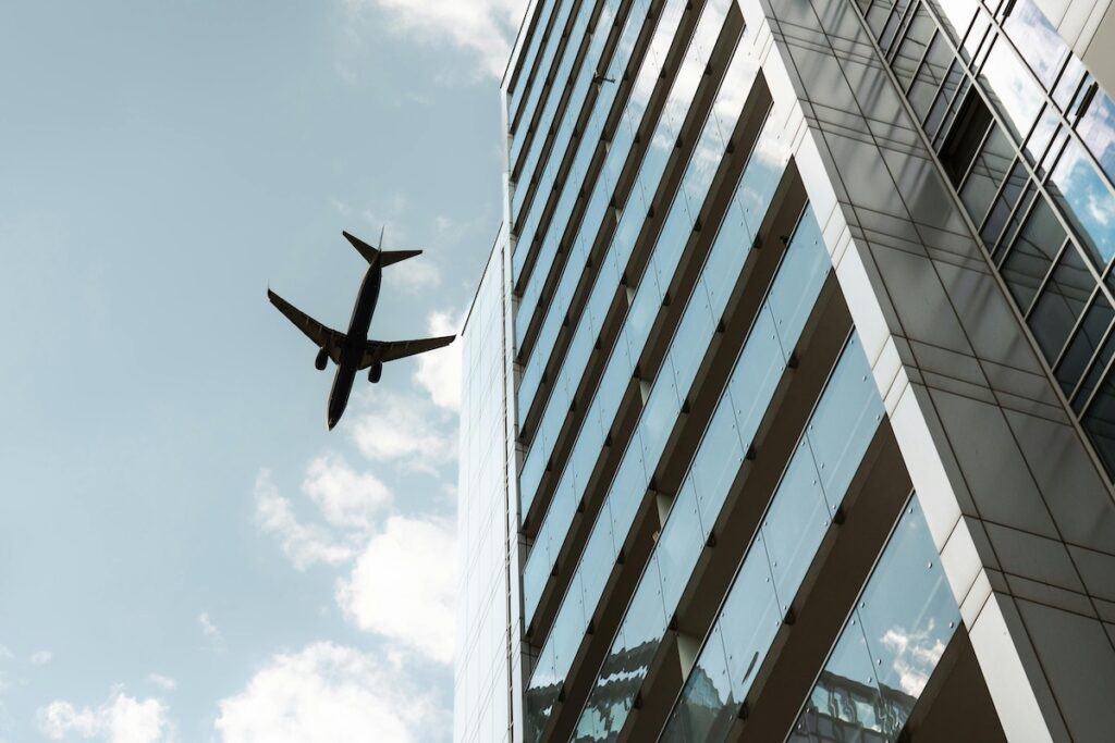 standing next to a tall building looking up at the sky and a plane flying overhead