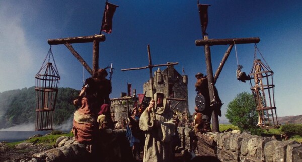 Film still from Highlander (1986) showing a hooded priest holding a cross made from sticks, walking across a bridge