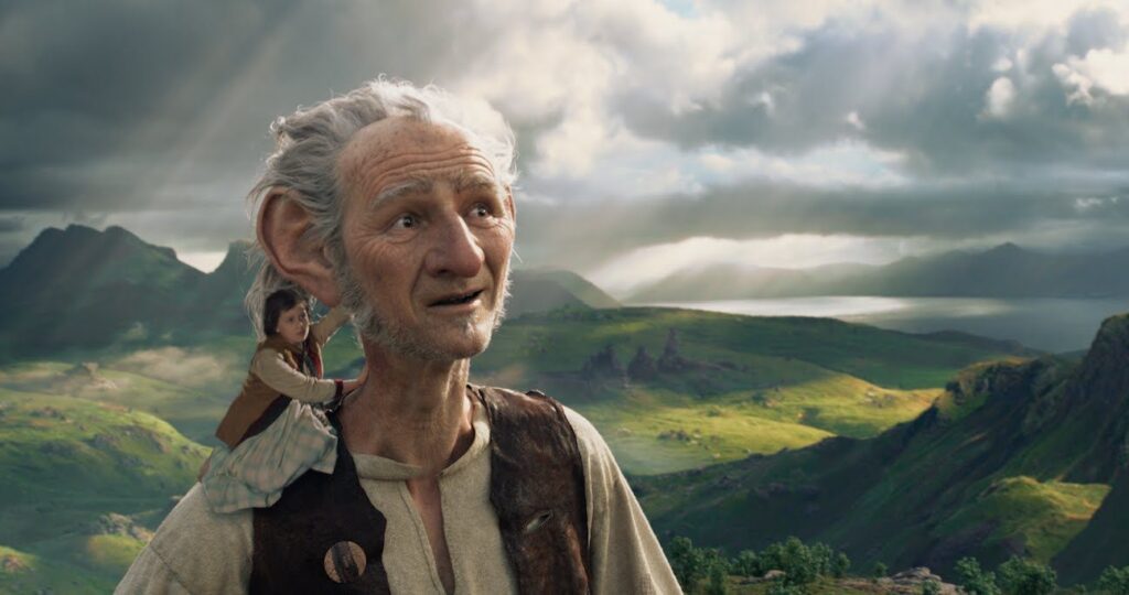 Film still from The BFG (2016) showing soph sat on top of the BFG's shoulder with green hills in the background