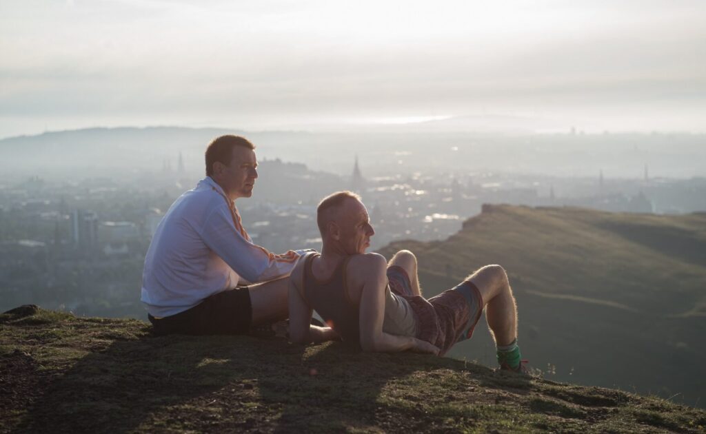 Film still from Trainspotting (1996) showing two men lying on a hill looking out at the Ediburgh skyline