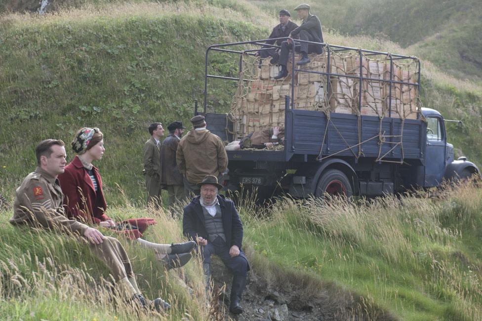 Film still from Whisky Galore! (2016) showing characters sitting on a grassy hilltop.